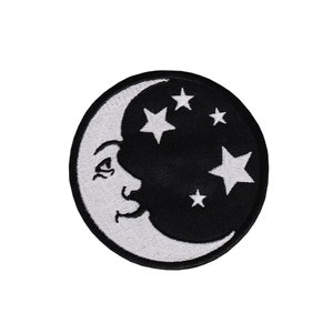 Könnte beinhalten: Ein weißer Halbmond mit einem Gesicht und fünf weißen Sternen auf schwarzem Hintergrund. Mond und Sterne sind auf einem schwarzen Stoffpatch gestickt.