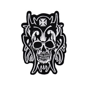 Könnte beinhalten: Schwarz-weiß gestickter Patch mit einem Schädel mit Hörnern und einem Kreuz. Der Schädel ist von einem dekorativen Design umgeben.