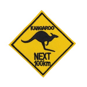 Op de afbeelding: Een geel, ruitvormig waarschuwingsbord met een zwarte rand en tekst. Het bord luidt "Kangaroo Next 100km" en toont een zwart silhouet van een springend kangoeroe.