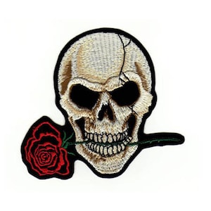 Könnte beinhalten: Gestickter Bügel-Patch mit einem weißen Totenkopf, der eine rote Rose im Mund hält. Der Totenkopf hat eine rissige Oberfläche und schwarze Augenhöhlen. Die Rose hat einen grünen Stiel und Blätter.