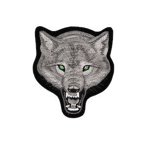 Könnte beinhalten: Gestickter Aufnäher mit dem Gesicht eines knurrenden Wolfes mit grünen Augen. Das Fell des Wolfes ist grau und der Hintergrund ist schwarz.