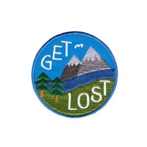 Könnte beinhalten: Ein blauer und grüner gestickter Aufnäher mit einer Bergkette und Bäumen. Der Text "GET LOST" ist in Weiß geschrieben.