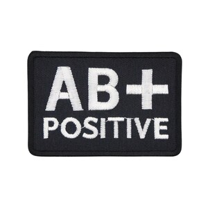 May include: A black rectangular patch with white embroidered text. The text reads "AB+ POSITIVE". The patch has a black border and is likely made of fabric. The text is in a bold, sans-serif font.