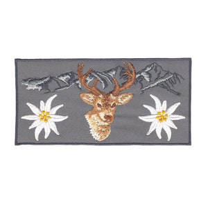 Könnte beinhalten: Gestickter Aufnäher mit einem braunen Hirschkopf, zwei weißen Edelweißblüten und einer grauen Bergkette auf grauem Hintergrund.