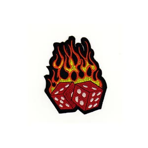 Könnte beinhalten: Zwei rote Würfel mit weißen Punkten stehen in Flammen. Die Flammen sind gelb und orange mit schwarzen Umrissen. Die Würfel sind auf einem schwarzen Stoff gestickt.