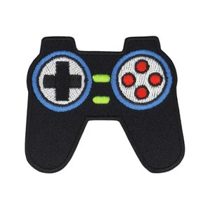 Könnte beinhalten: Schwarzer gestickter Patch eines Videospiel-Controllers mit blauen und roten Knöpfen. Der Controller hat ein weißes Kreuz auf dem linken Knopf und vier rote Kreise auf dem rechten Knopf.