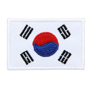 Puede incluir: Un parche blanco bordado con la bandera de Corea del Sur. La bandera presenta un círculo rojo y azul con cuatro líneas horizontales negras sobre un fondo blanco.