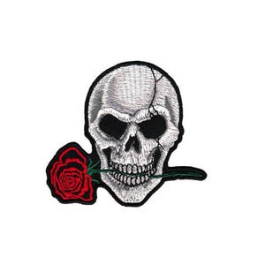 Könnte beinhalten: Ein weißer Schädel mit einem rissigen Gesicht hält eine einzelne rote Rose in seinen Zähnen. Der Schädel ist in weißer Farbe auf einem schwarzen Hintergrund gestickt.