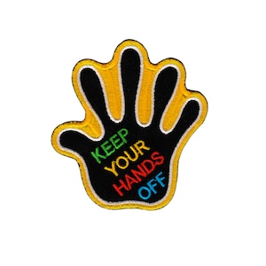 Op de afbeelding: Een gele handvormige patch met zwarte stiksels en de woorden "KEEP YOUR HANDS OFF" in kleurrijke borduurwerk.