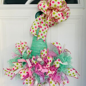 Puede incluir: Una corona verde, blanca y rosa con un gran lazo de lunares. La corona está hecha de malla y cinta y tiene una variedad de texturas.