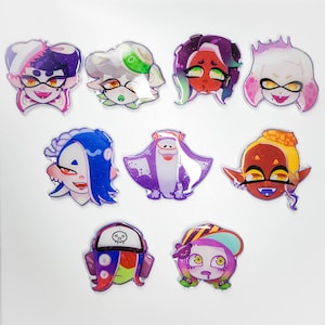 May include: A set of ten colorful cartoon character pins featuring characters from the video game Splatoon. The pins are made of clear plastic with a holographic finish. The characters are all different colors and have different expressions on their faces.