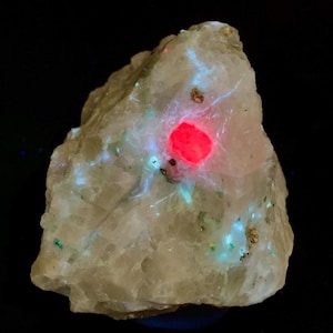 Puede incluir: Una roca rugosa de color claro con un centro rojo vibrante y acentos azules y verdes brillantes. La superficie de la roca tiene una apariencia texturizada, con pequeñas motas marrones y verdes dispersas. El fondo es negro sólido.
