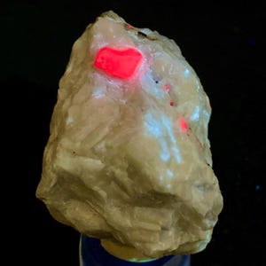 May include: A beige-coloured mineral specimen with a bright, glowing red inclusion. The rock has a textured surface and is displayed against a dark background. Small red and white glowing spots are visible on the surface.