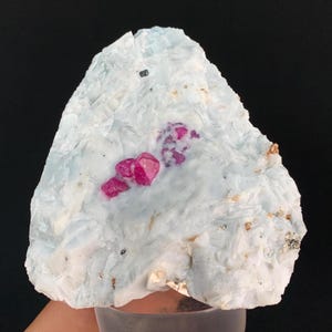 Puede incluir: Un espécimen mineral en bruto que presenta una matriz cristalina de color azul pálido y gélido con prominentes racimos de cristales de color magenta vibrante incrustados en la superficie. La estructura es irregular y dentada, mostrando un contraste entre la base azul claro y las inclusiones minerales de color rosa intenso.