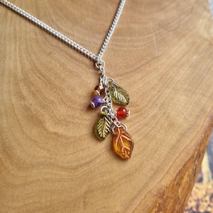 Może przedstawiać: Srebrny łańcuszek z zawieszką przedstawiającą wiele szklanych liści w odcieniach zieleni, pomarańczy i bursztynu. Zawieszka zawiera również małe okrągłe koraliki w kolorze fioletowym, pomarańczowym i czerwonym.