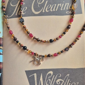 Może przedstawiać: Wielowarstwowy naszyjnik z zawieszką w kształcie konia. Naszyjnik posiada mieszankę brązowych, czerwonych, czarnych i bursztynowych koralików. Zawieszka to koń w kolorze srebrnym. Naszyjnik jest prezentowany na książce z tytułem "The Clearing" i "Wolf Alice".