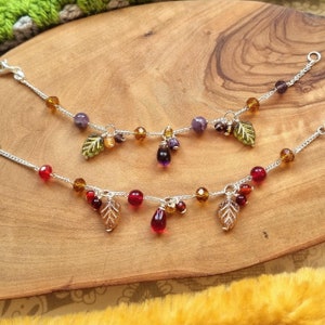 May include: Two silver chain bracelets with dangling charms. The top bracelet has purple, amber, and green beads with a green leaf charm. The bottom bracelet has red, amber, and green beads with a gold leaf charm.