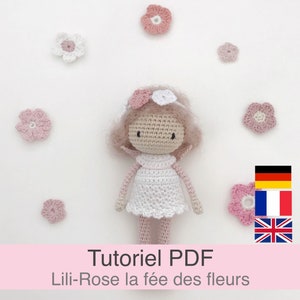 Może przedstawiać: Lalka szydełkowa z różowymi i białymi kwiatami we włosach, ubrana w białą sukienkę z różowymi lamówkami. Lalka jest otoczona różowymi kwiatami. Tekst "Tutoriel PDF Lilli-Rose la fée des fleurs" znajduje się na dole obrazu. Widoczne są również flagi Niemiec, Francji i Wielkiej Brytanii.