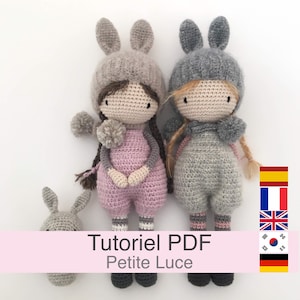 Könnte beinhalten: Zwei handgefertigte Häkelpuppen mit Hasenohrenhüten und Overalls, eine rosa und eine graue. Ein kleiner grauer Hasenkopf ist ebenfalls vorhanden. Die Puppen haben Garnhaare und Bommelakzente. Text auf einem rosa Banner lautet "Tutoriel PDF Petite Luce."