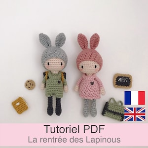 Peut inclure: Deux poupées de lapin en crochet habillées en tenue scolaire. La poupée de gauche porte une tenue verte et un sac à dos. La poupée de droite porte une tenue rose. Les deux poupées ont des oreilles de lapin grises et tiennent un petit cœur. Les poupées sont assises sur une surface blanche avec un tableau noir, un livre et un biscuit.