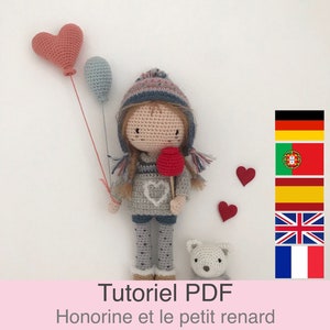 Może przedstawiać: Lalka szydełkowa z lizakiem w kształcie czerwonego serca, szarym i białym swetrem oraz szarą i białą spódnicą w kropki. Lalka trzyma lizak w kształcie czerwonego serca i ma szary i biały kapelusz. Lalka stoi na białym tle z dwoma czerwonymi sercami i białym szydełkowym psem.