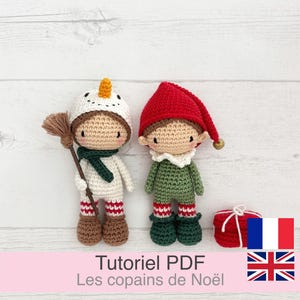 Op de afbeelding: Twee handgemaakte gehaakte poppen in kerstkleding. Eén pop is gekleed als een sneeuwpop met een bezem, de andere als een elf met een rode hoed. Beide poppen hebben bruin haar en staan op een witte houten ondergrond.