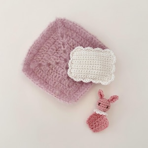 May include: A collection of handmade crochet items in soft pastel colors. The image features a square, fluffy, mauve-colored piece, a white rectangular piece with a scalloped edge, and a small pink bunny with a white collar.
