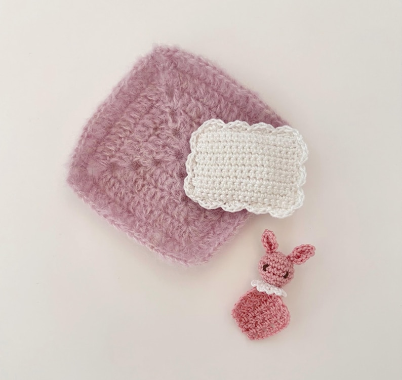 May include: A collection of handmade crochet items in soft pastel colors. The image features a square, fluffy, mauve-colored piece, a white rectangular piece with a scalloped edge, and a small pink bunny with a white collar.
