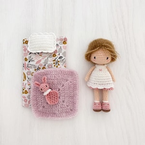 May include: A handmade crochet doll with brown hair and a white dress with pink trim. A small crochet bed set includes a floral patterned blanket, a white pillow, and a pink square blanket with a crochet bunny. The doll is approximately 5 inches tall.