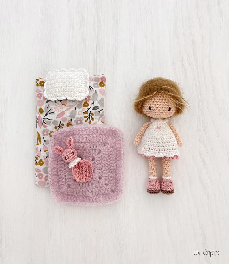 May include: A handmade crochet doll with brown hair and a white dress with pink trim. A small crochet bed set includes a floral patterned blanket, a white pillow, and a pink square blanket with a crochet bunny. The doll is approximately 5 inches tall.