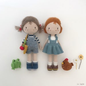 Puede incluir: Dos mu&ntilde;ecas de crochet con cabello casta&ntilde;o, que llevan monos y vestidos azules, de pie junto a una rana verde y un pollo marr&oacute;n. Las mu&ntilde;ecas tienen ojos marrones y mejillas sonrosadas.