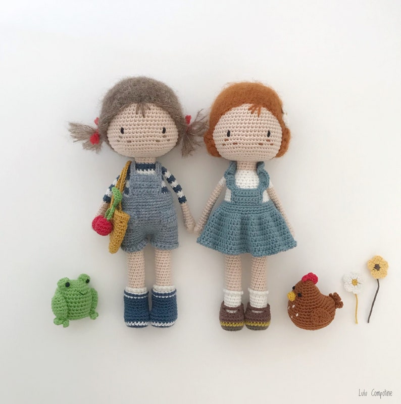 Puede incluir: Dos mu&ntilde;ecas de crochet con cabello casta&ntilde;o, que llevan monos y vestidos azules, de pie junto a una rana verde y un pollo marr&oacute;n. Las mu&ntilde;ecas tienen ojos marrones y mejillas sonrosadas.
