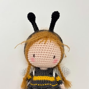 May include: A handmade crochet doll resembling a bee. The doll features a light pink face, red braided hair, and black antennae. The dress is black with yellow stripes, and it has small, silver wings.