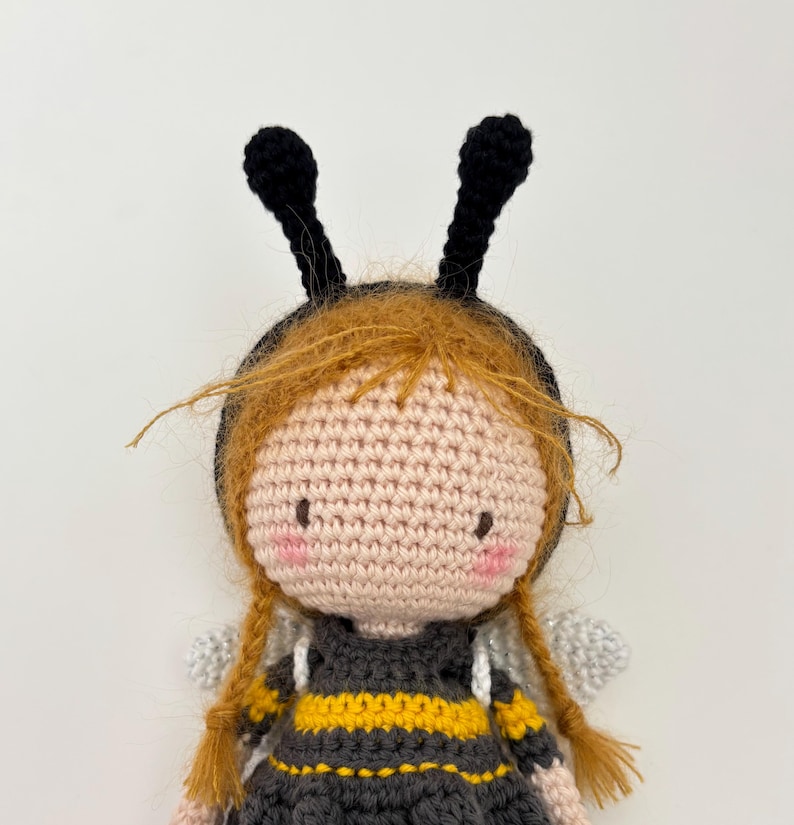 May include: A handmade crochet doll resembling a bee. The doll features a light pink face, red braided hair, and black antennae. The dress is black with yellow stripes, and it has small, silver wings.