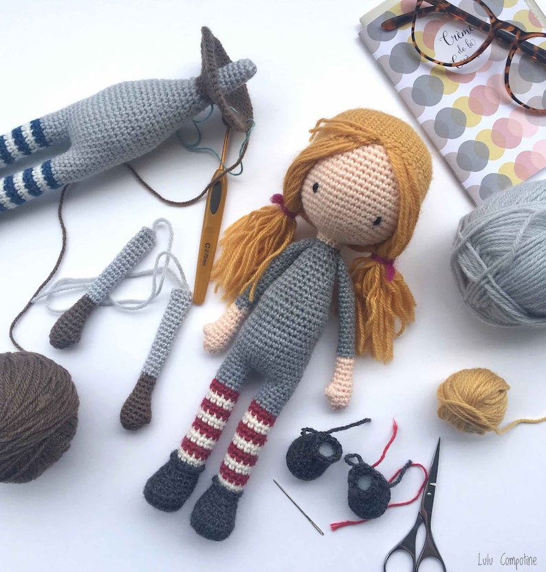 Peut inclure: Une poup&eacute;e en crochet avec des cheveux blonds, une robe grise, un legging ray&eacute; rouge et blanc et des chaussures grises. La poup&eacute;e est entour&eacute;e de laine, de crochets et d'autres parties de poup&eacute;e.