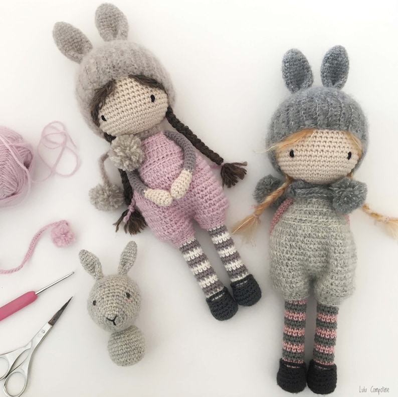 Pode incluir: Bonecas de croch&ecirc; feitas &agrave; m&atilde;o com chap&eacute;us de orelhas de coelho. Uma boneca usa um macac&atilde;o rosa com pernas listradas cinza e branco, a outra usa um macac&atilde;o cinza com pernas listradas rosa e cinza. Uma cabe&ccedil;a de coelho cinza e fios tamb&eacute;m s&atilde;o vis&iacute;veis.