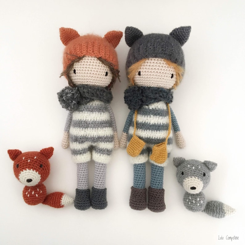 Op de afbeelding: Twee gehaakte poppen met gestreepte outfits en dierenmutsen, een vos en een wolf, met bijpassende gehaakte dierenbegeleiders. De poppen dragen sjaals en hebben bruine ogen.