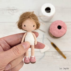 May include: A handmade crochet doll with brown hair, a white dress, and pink shoes. The doll is held in a hand, with spools of white and pink yarn, a crochet hook, and the artist's signature in the background. The doll is approximately 3 inches tall.