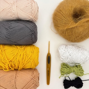 May include: An overhead view of crochet supplies, including yarn and a hook. The yarn is in shades of pink, grey, yellow, and tan. There are also smaller bundles of white, green, and black yarn, and a golden crochet hook, all on a white background.