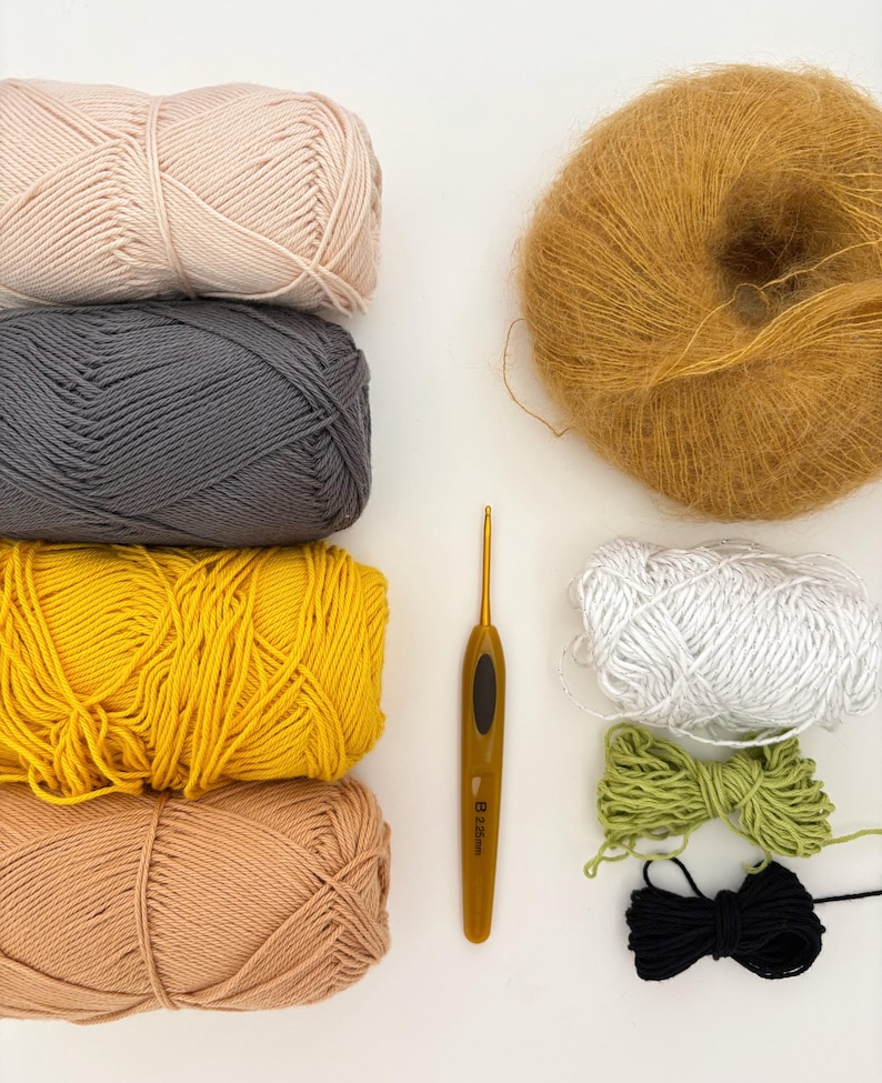 May include: An overhead view of crochet supplies, including yarn and a hook. The yarn is in shades of pink, grey, yellow, and tan. There are also smaller bundles of white, green, and black yarn, and a golden crochet hook, all on a white background.
