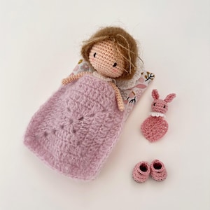May include: A handmade crochet doll with brown hair, lying in a pink blanket on a floral patterned pillow. Includes a pink bunny toy and a pair of pink crochet shoes. The doll has a white collar.
