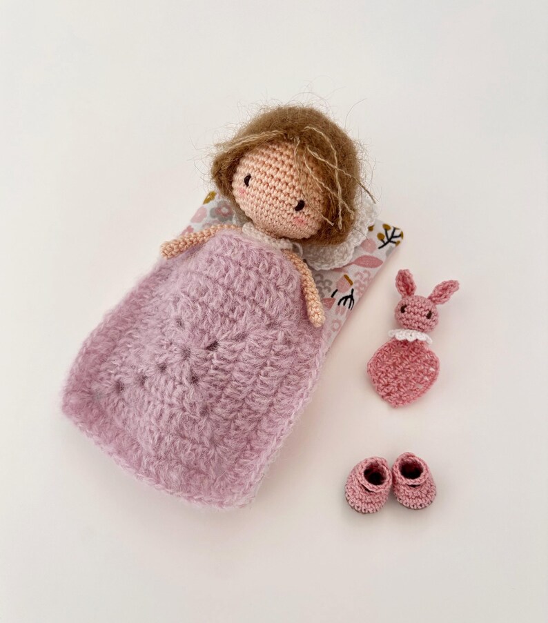May include: A handmade crochet doll with brown hair, lying in a pink blanket on a floral patterned pillow. Includes a pink bunny toy and a pair of pink crochet shoes. The doll has a white collar.