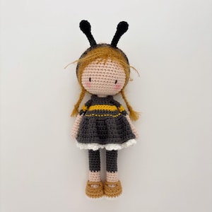 May include: A handcrafted crochet doll resembling a bee. The doll features a fair complexion, ginger braids, and a black and yellow striped dress. It is adorned with a black headband with antennae and brown shoes. The background is plain white.