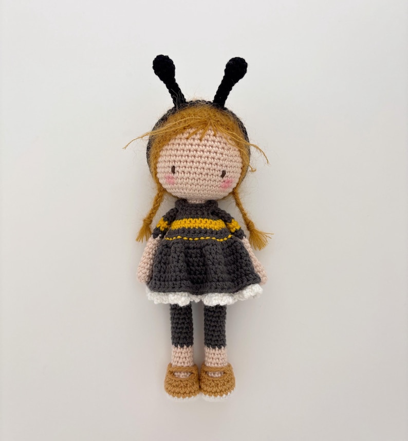 May include: A handcrafted crochet doll resembling a bee. The doll features a fair complexion, ginger braids, and a black and yellow striped dress. It is adorned with a black headband with antennae and brown shoes. The background is plain white.