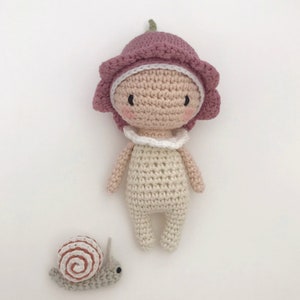 Puede incluir: Una mu&ntilde;eca de ganchillo hecha a mano con un sombrero en forma de flor en tonos rosas y un cuello blanco. La mu&ntilde;eca tiene un tono de piel claro y un peque&ntilde;o caracol de ganchillo al lado. La mu&ntilde;eca mide unos 15 cm de alto.