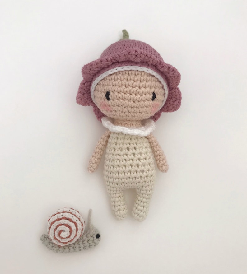 Puede incluir: Una mu&ntilde;eca de ganchillo hecha a mano con un sombrero en forma de flor en tonos rosas y un cuello blanco. La mu&ntilde;eca tiene un tono de piel claro y un peque&ntilde;o caracol de ganchillo al lado. La mu&ntilde;eca mide unos 15 cm de alto.