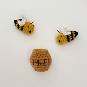 May include: A crocheted bee and honey pot set. The bees are yellow with black stripes and white wings. The honey pot is brown and has the word "MIE" embroidered on it. Ideal for a nursery or as a gift, this set is handmade.