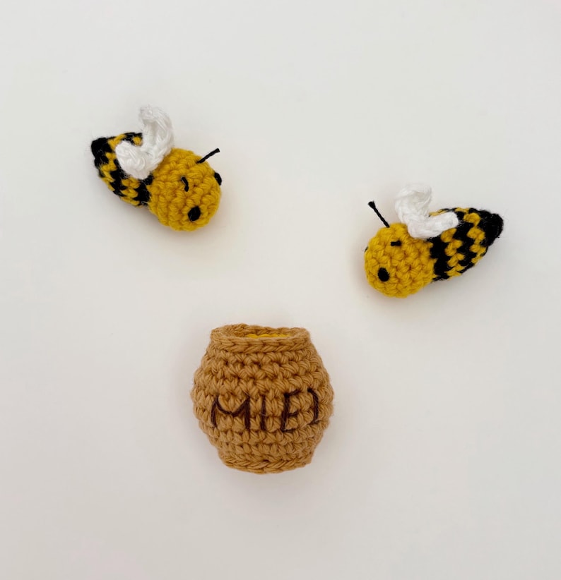 May include: A crocheted bee and honey pot set. The bees are yellow with black stripes and white wings. The honey pot is brown and has the word "MIE" embroidered on it. Ideal for a nursery or as a gift, this set is handmade.