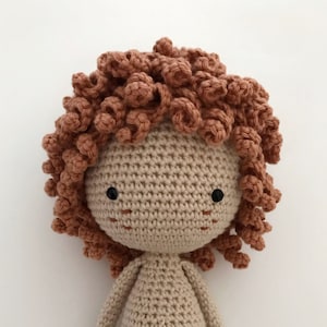 K&ouml;nnte beinhalten: Eine geh&auml;kelte Puppe mit einem beigefarbenen K&ouml;rper und braunen, lockigen Haaren. Die Puppe hat schwarze Augen und ein einfaches, gesticktes L&auml;cheln.