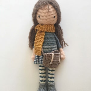 Peut inclure: Une poup&eacute;e en crochet avec des cheveux bruns, une &eacute;charpe jaune, un pull ray&eacute; gris et blanc, une jupe &agrave; carreaux, des leggings ray&eacute;s et des chaussures brunes. La poup&eacute;e tient un sac en crochet brun.